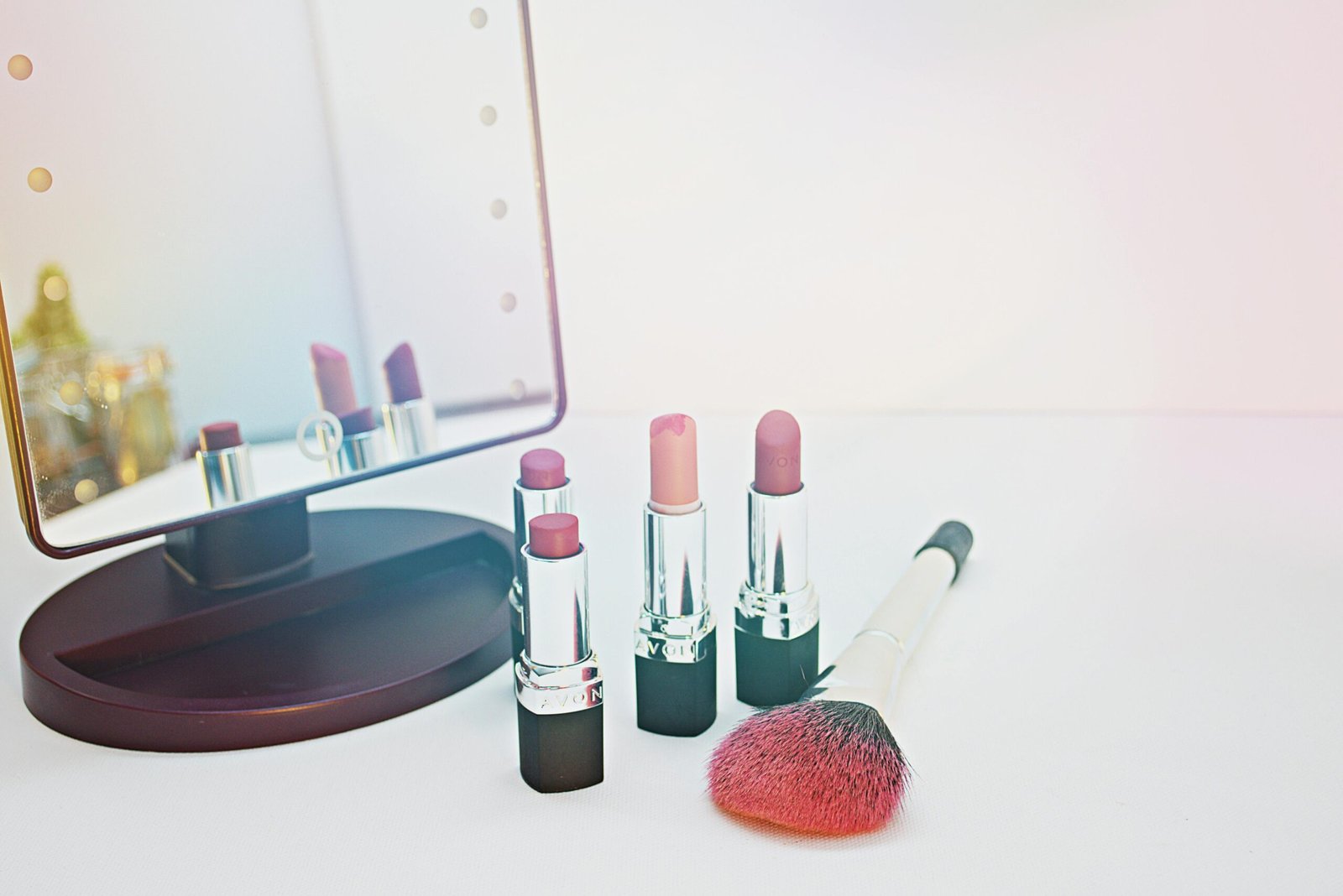 A bright, clean shot of four lipsticks in shades of pink and mauve, standing upright on a white surface next to a large pink-tipped makeup brush. A light-up vanity mirror sits to the left, reflecting the lipsticks.