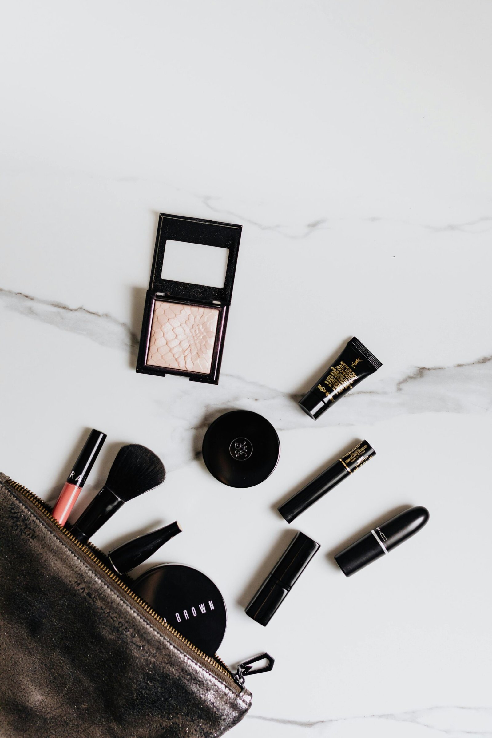 A top-down view of high-end makeup products scattered on a white marble surface next to an open black makeup bag. Visible items include an Yves Saint Laurent foundation tube, a MAC lipstick, a Bobbi Brown compact, and various brushes and eyeliners.